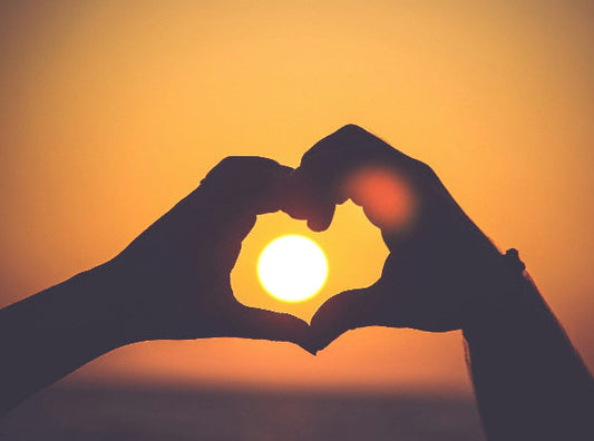 Two hands making a heart shape against a sunset sky.